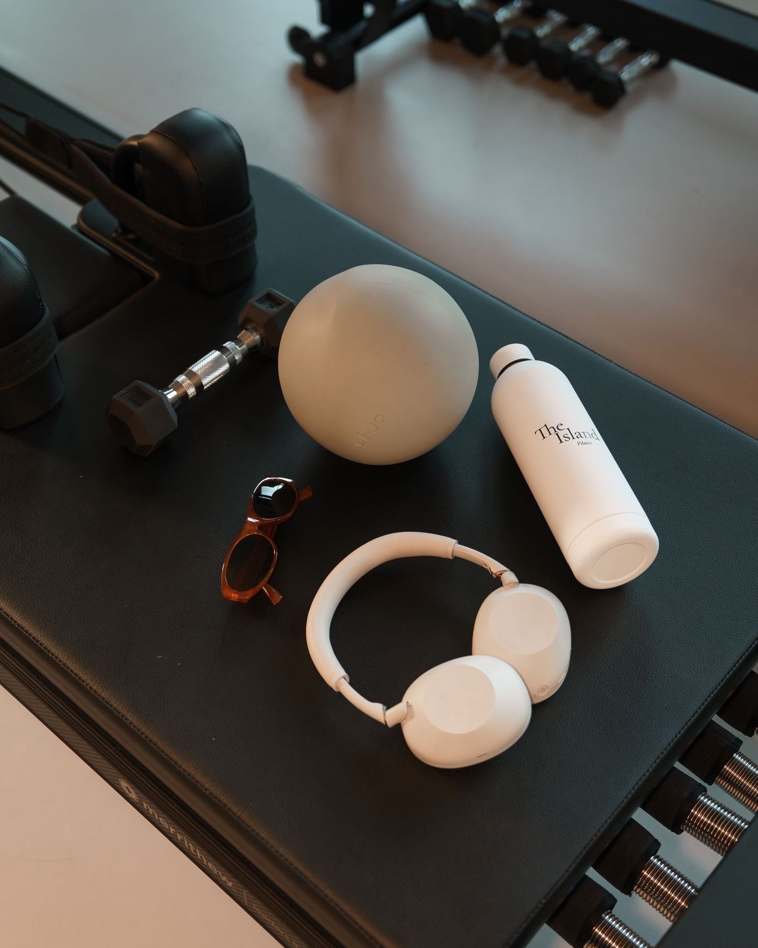 Flat lay of white headphones, water bottle, ball, sunglasses, and gym equipment on a dark surface.