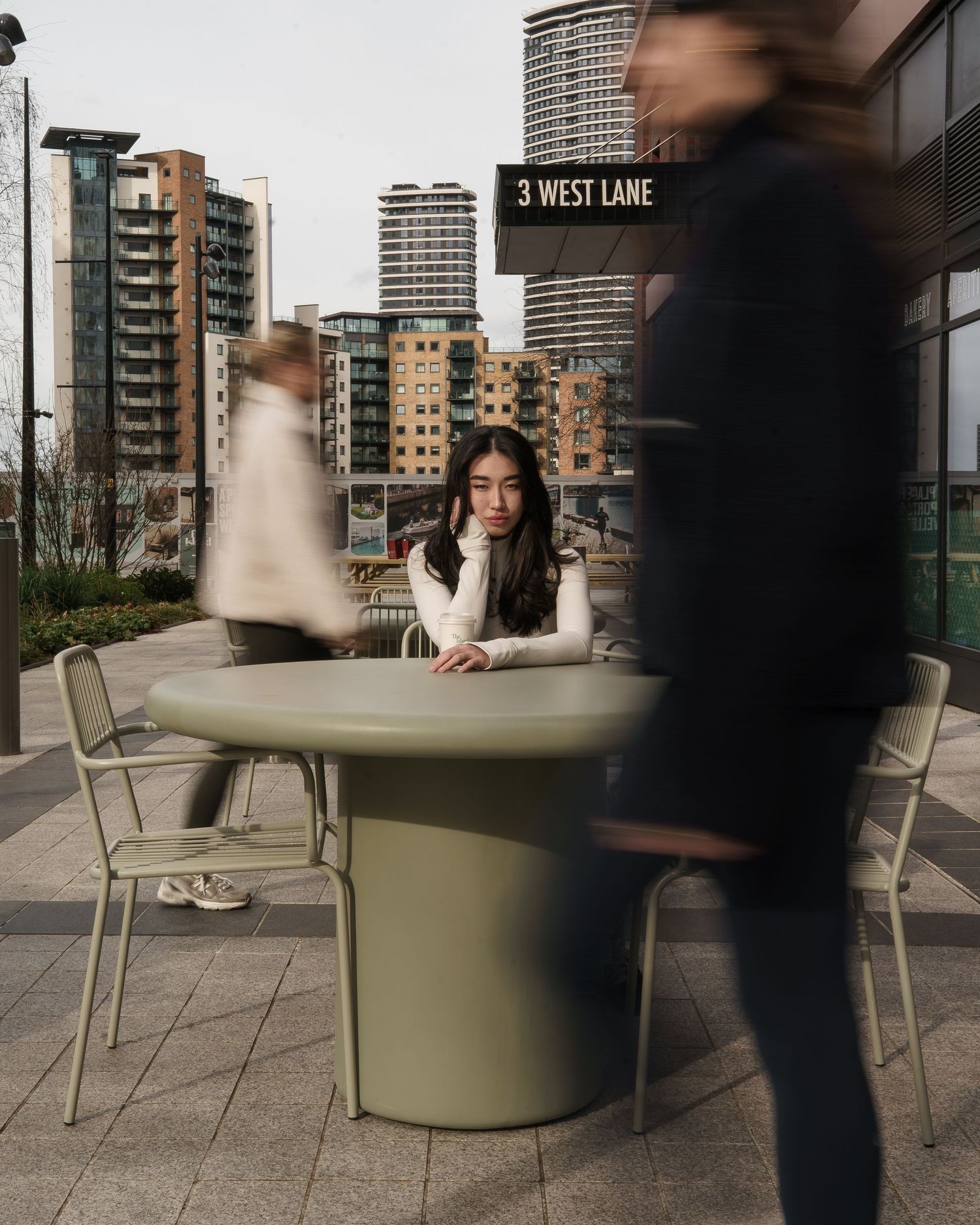 Three young women walking on a city sidewalk carrying bags and coffee cups near a brick building.
