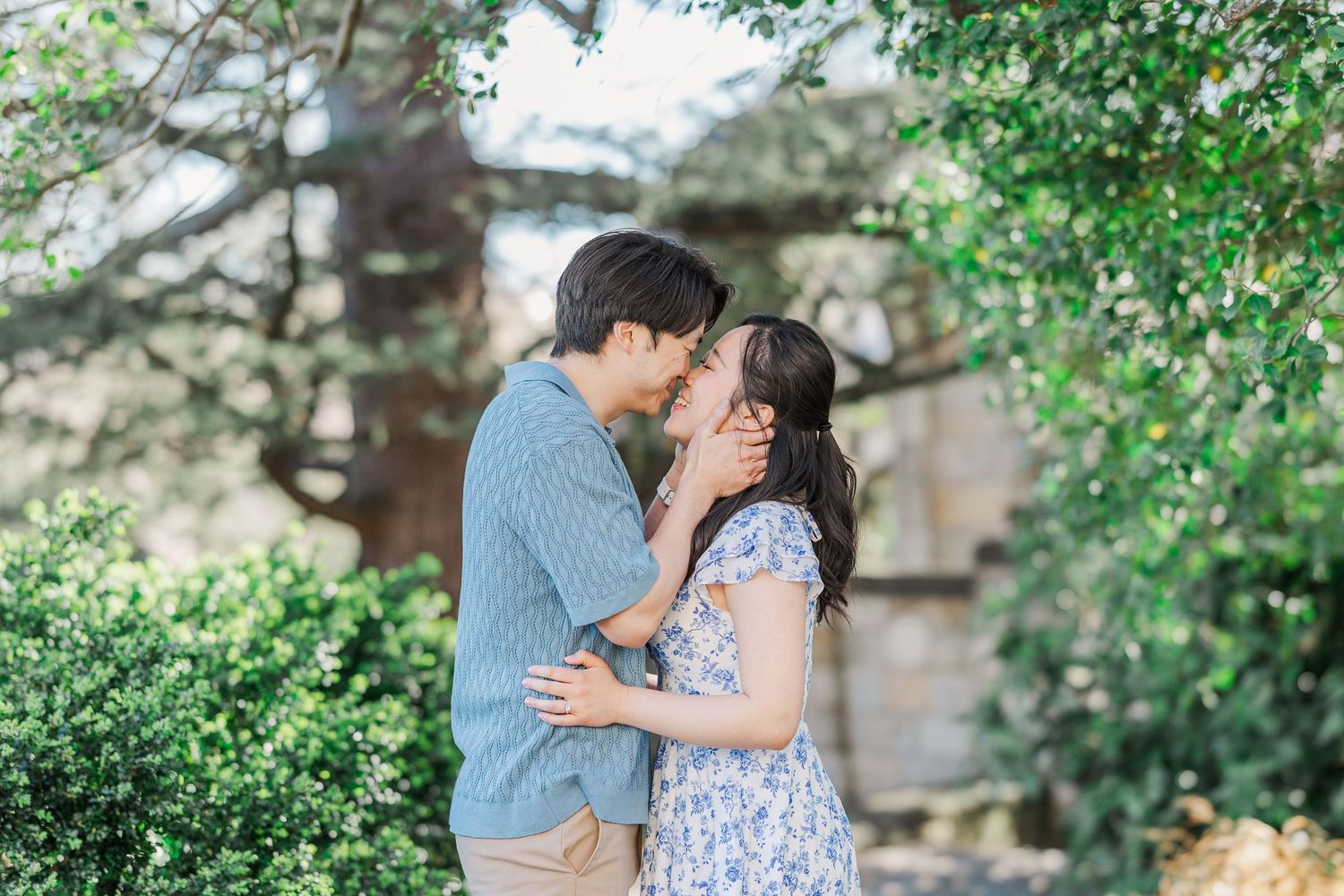 Couple sharing a romantic almost-kiss moment outdoors surrounded by lush green bushes and trees.