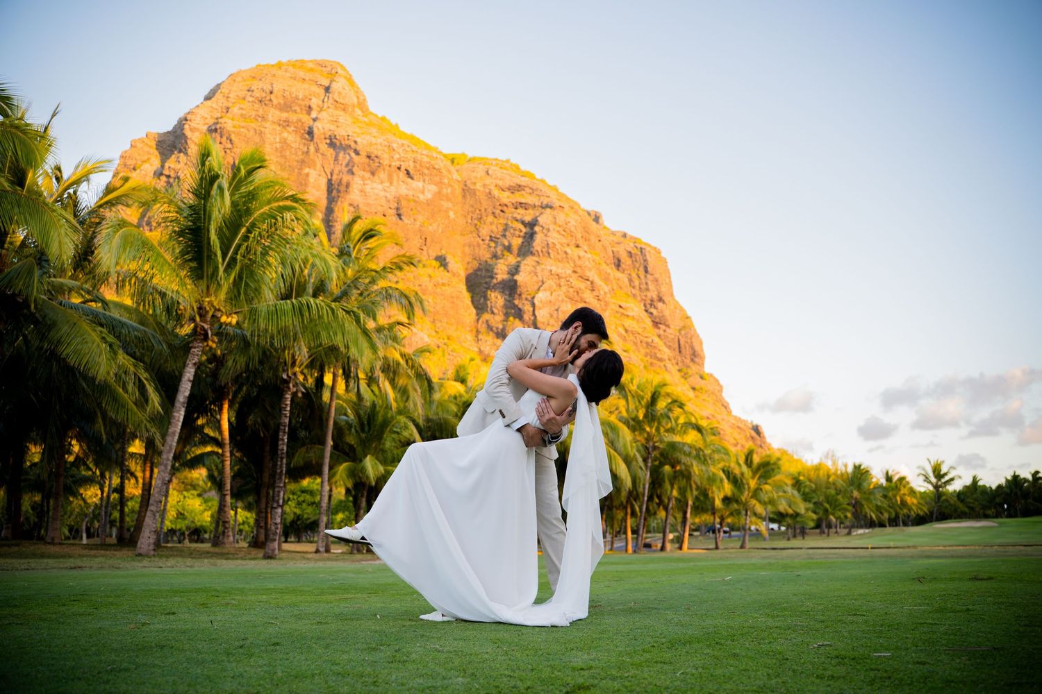 Couple sharing a romantic dip kiss on lush green lawn with tropical palm trees and dramatic mountain at golden hour.