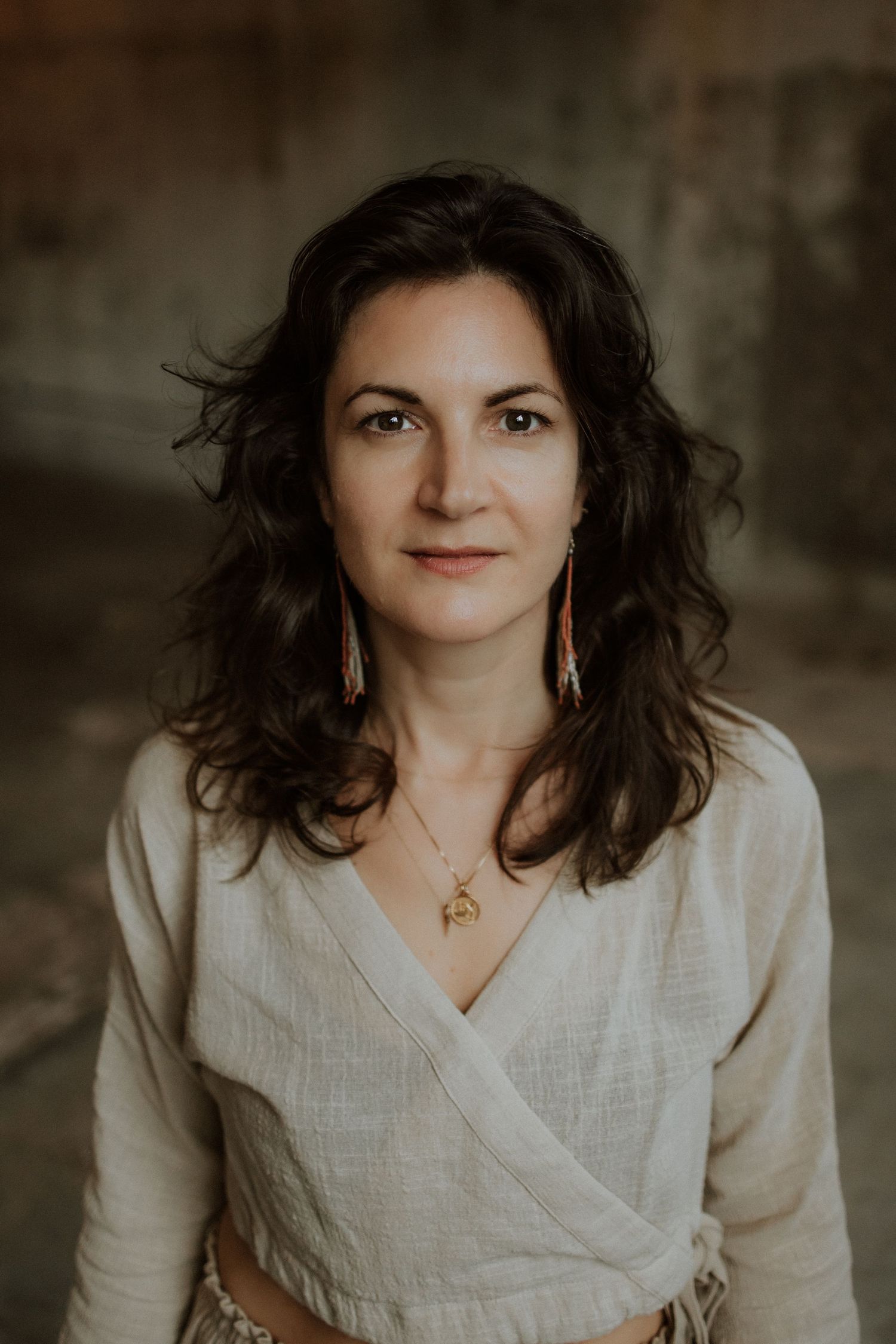 Woman with dark curly hair wearing a beige wrap top and gold jewelry, posing for a portrait photo.