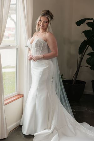 Smiling bride in strapless beaded mermaid wedding gown with veil stands by window in elegant bridal suite.