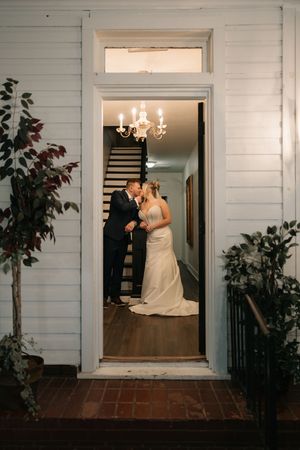 Bride and groom share a romantic kiss in a warmly lit doorway of a white wooden home with a chandelier above.