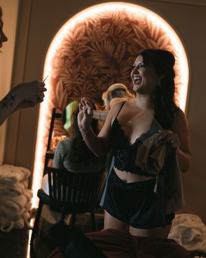 Woman in black lace lingerie laughing joyfully in a warmly lit room with an illuminated arched mirror.