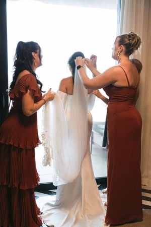 Two bridesmaids in rust dresses help a bride with her veil and wedding gown near a bright window.