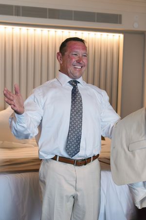 Smiling middle-aged man in white shirt and tie gesturing welcomingly in a hotel room.