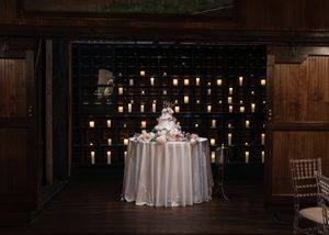 Elegant wedding sweetheart table with white linen, floral centerpiece, and glowing candle wall backdrop in a dark venue.