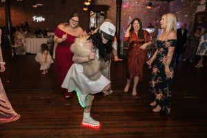 Woman dancing in light-up shoes and trucker hat at a wedding reception while guests watch and smile.
