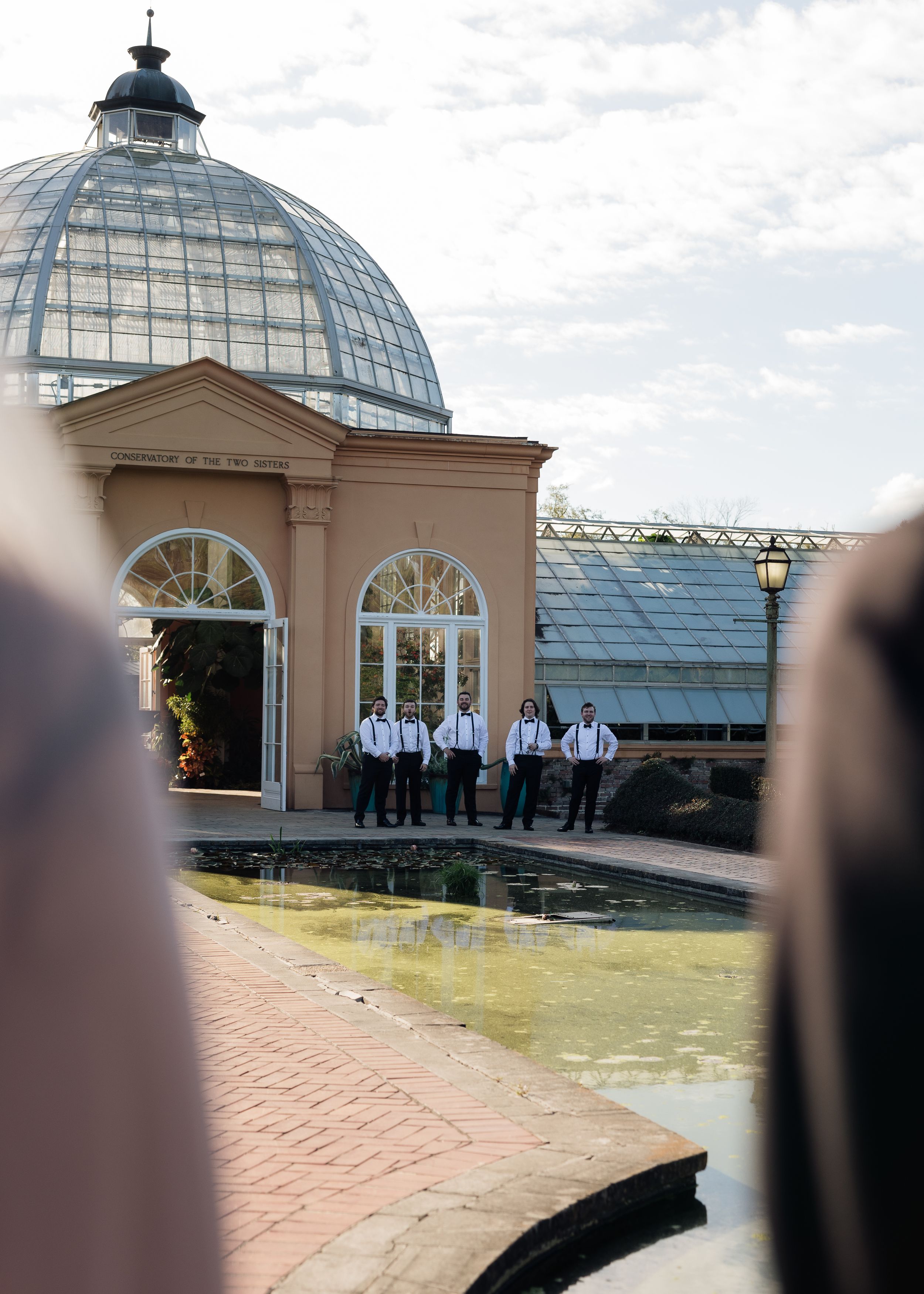 Groomsmen in white shirts and suspenders stand near a reflecting pool outside an elegant domed greenhouse venue.