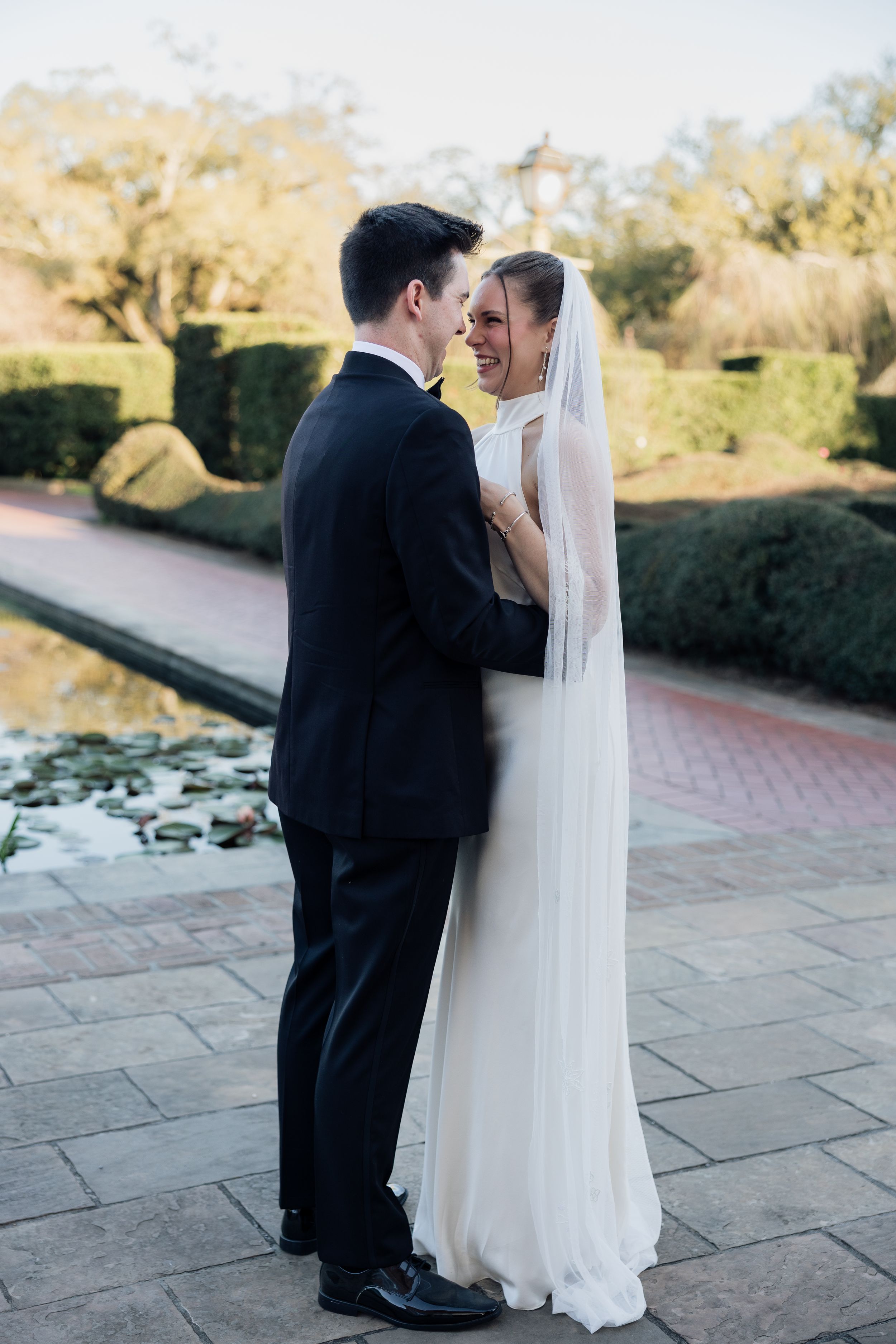 Bride and groom share a romantic moment in a formal garden, she in a flowing white gown with veil, he in a black tuxedo.