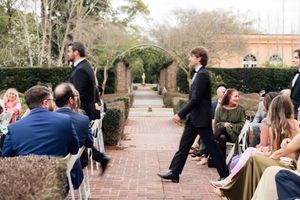 Two groomsmen walk down a brick aisle at an outdoor garden wedding ceremony with guests seated on both sides.
