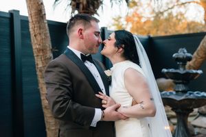 A bride and groom share a romantic kiss outdoors during their wedding ceremony, with lush greenery in the background.