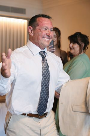 Smiling man in white shirt and tie gesturing at a formal event with guests in background.