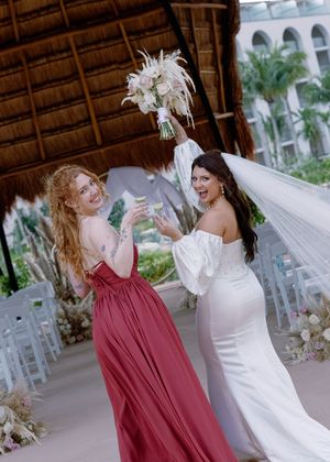 Bride in white gown raises bouquet with bridesmaid in red dress at tropical wedding ceremony.