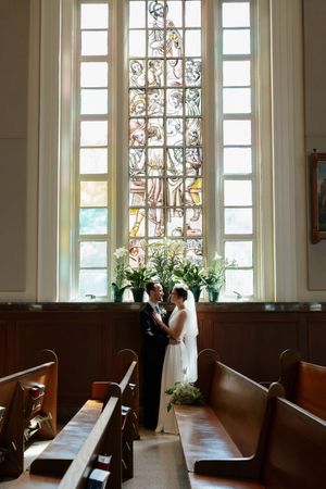 Bride and groom embrace before a stunning stained glass window in a historic church with wooden pews.