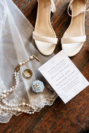 Bridal flat lay featuring white heels, pearl necklace, wedding rings in blue box, veil, and elegant invitation on wood surface.