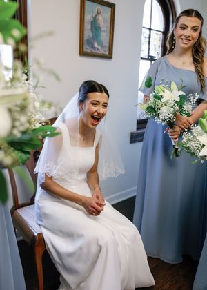 Laughing bride in white gown and veil sits beside smiling bridesmaid in blue dress holding white floral bouquet in church.