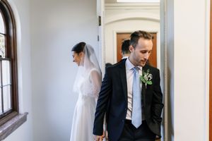 Bride in white gown and veil holds hands with groom in dark suit and boutonniere walking through doorway.