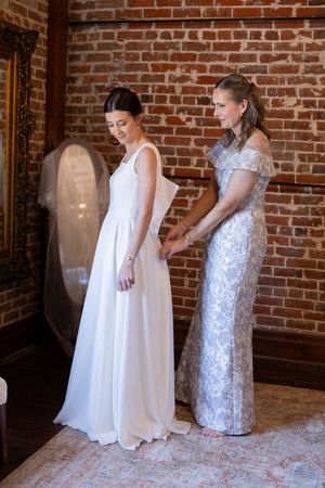 Bride in white gown holds hands with woman in silver lace dress near brick wall during wedding preparation.