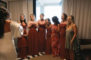 Bride reveals wedding dress to excited bridesmaids in rust-colored gowns, capturing joyful reactions during bridal party getting ready.