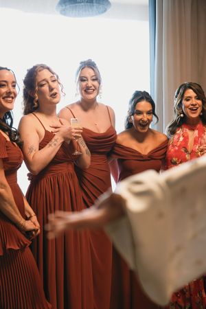 Bridesmaids in rust-colored gowns react with excitement as bride reveals wedding dress in bright hotel room.