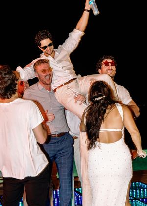 Group of friends celebrating at a party, man lifted on shoulders raising drink, woman in white backless dress.