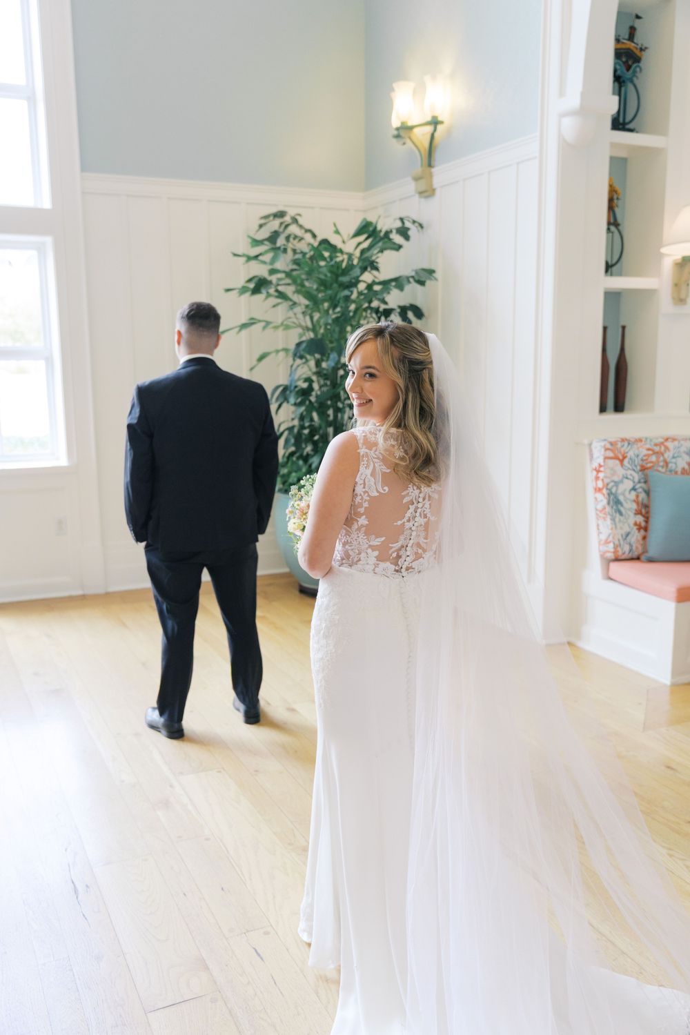 Bride in lace wedding gown with veil looks back during first look moment with groom indoors.