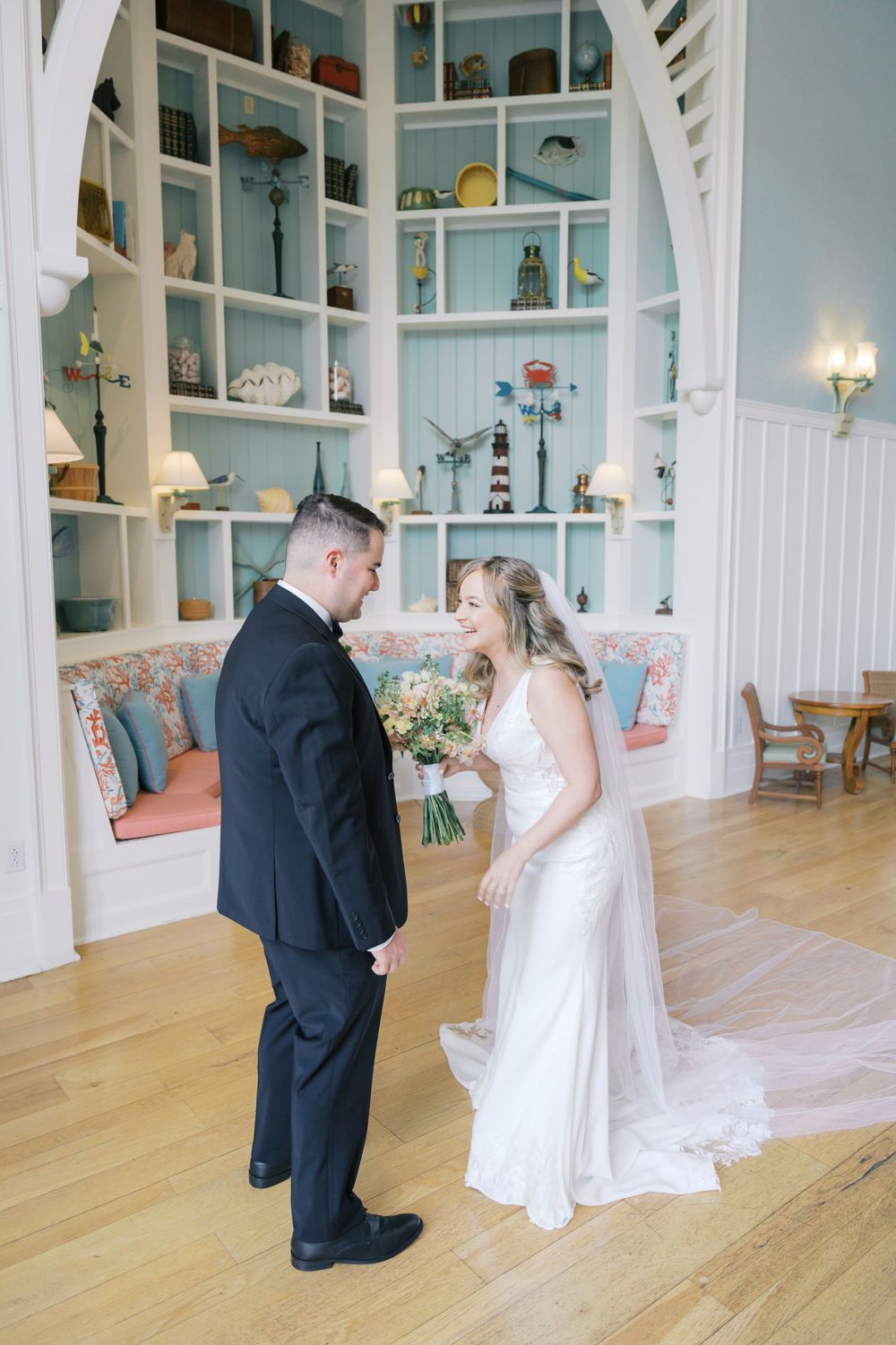 Smiling bride in a flowing white gown holds a bouquet while facing her groom in a navy suit in a charming coastal interior.