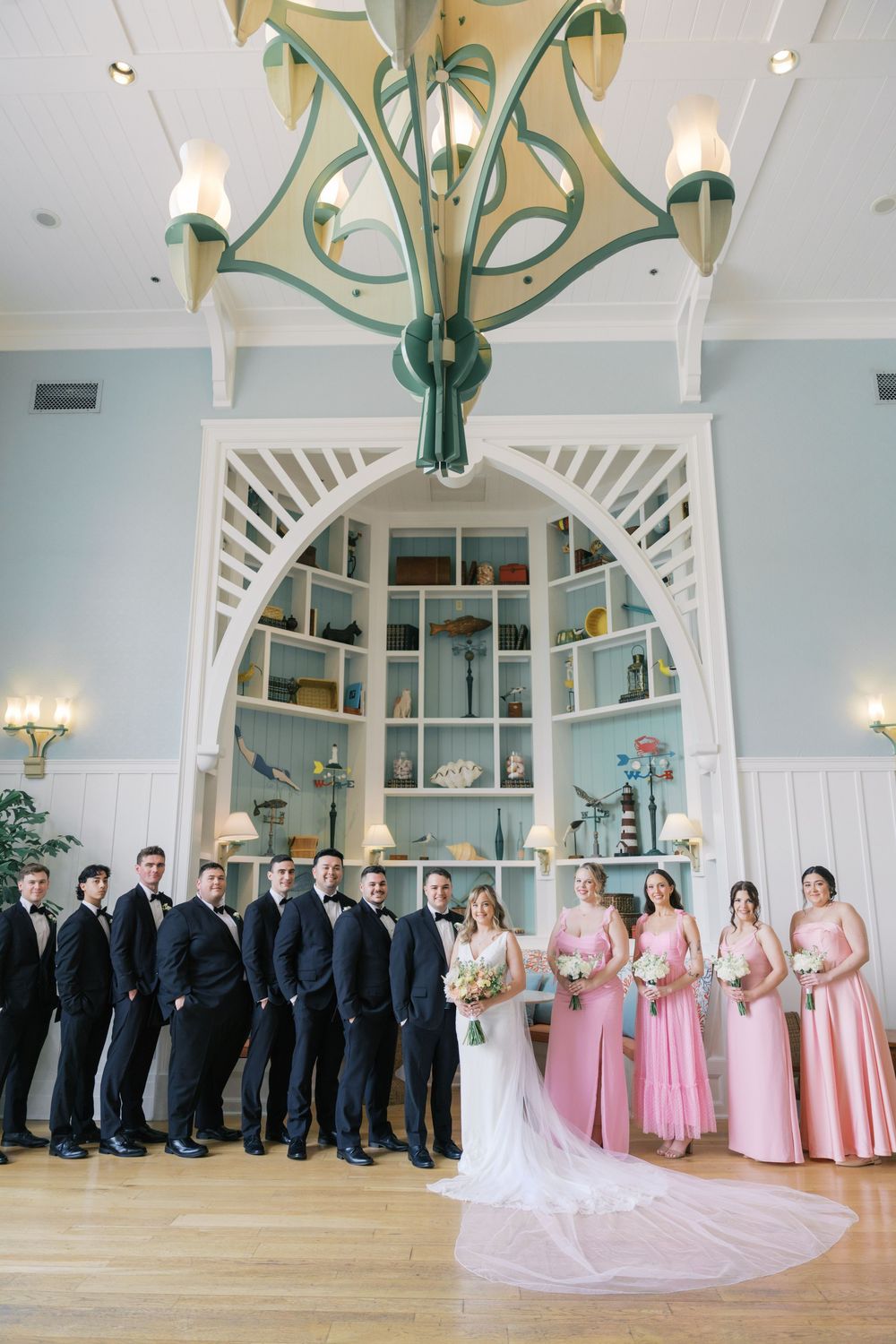 Wedding party poses under ornate chandelier with groomsmen in tuxedos and bridesmaids in pink gowns.