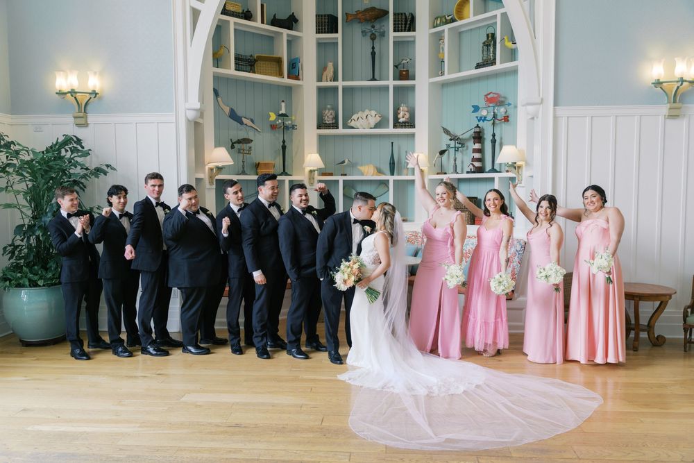 Wedding party poses in elegant venue, groomsmen in black tuxedos and bridesmaids in pink gowns flanking bride and groom.