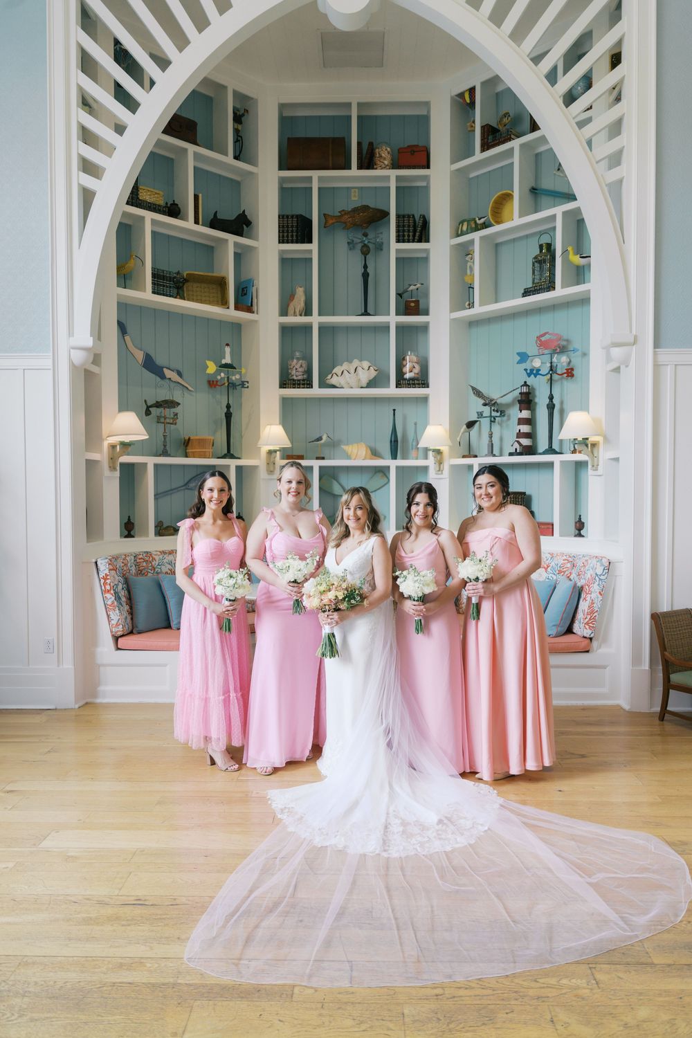 Bride in white gown poses with four bridesmaids in pink dresses inside a coastal-themed venue with blue shelving.