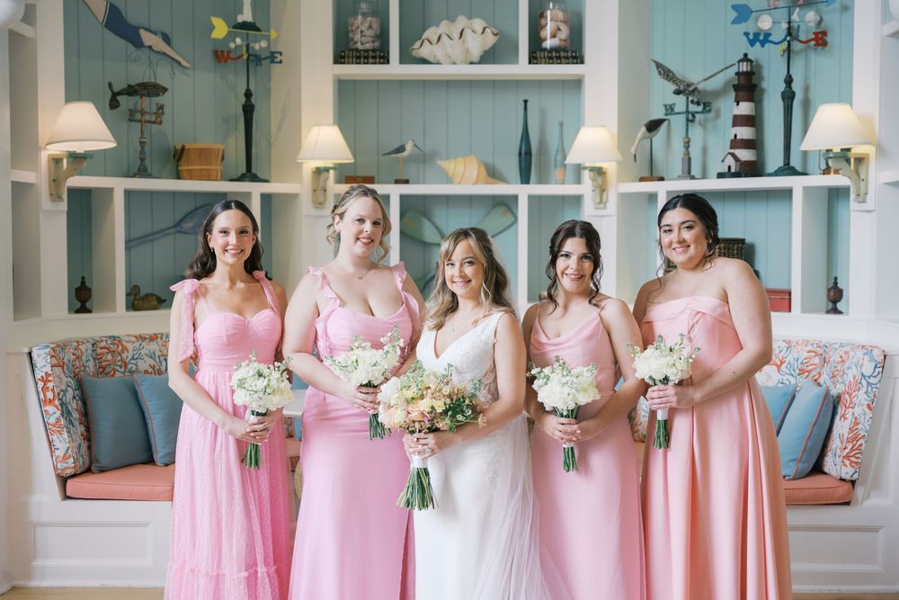 Bride with three bridesmaids in pink gowns holding white bouquets in a coastal-themed room.