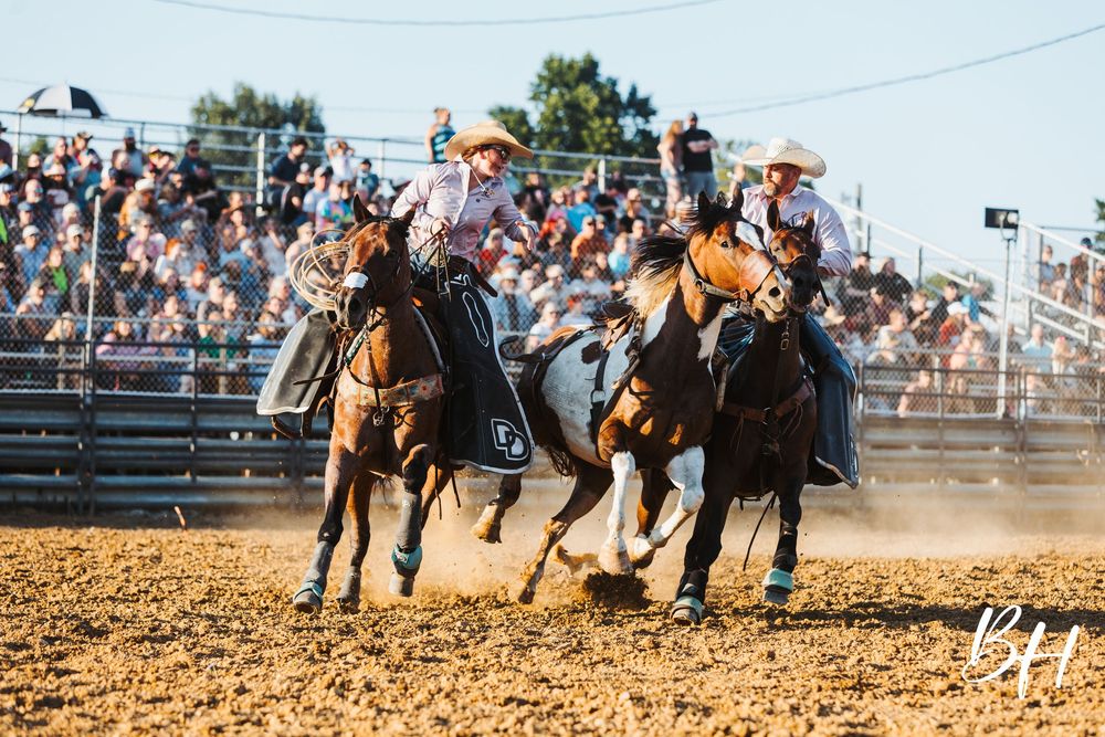 Cowboys on horseback compete in a rodeo event, kicking up dirt before a packed grandstand crowd.