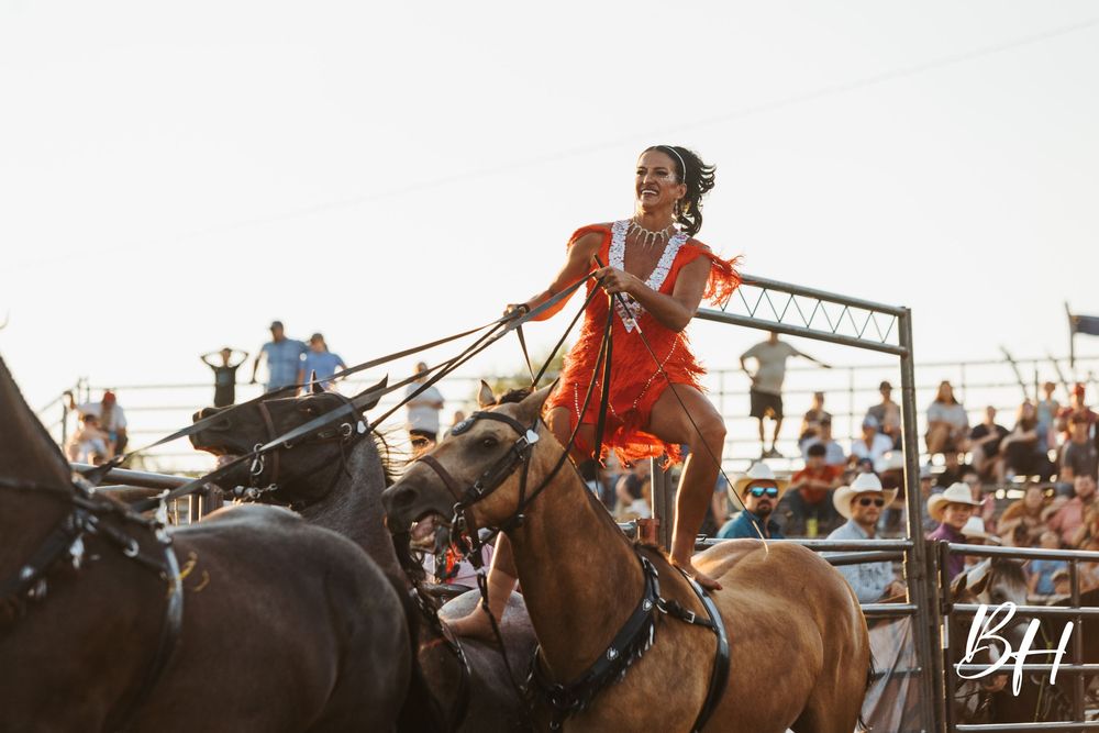 A woman in an orange dress rides horses at a rodeo event with a crowd of spectators in the background.
