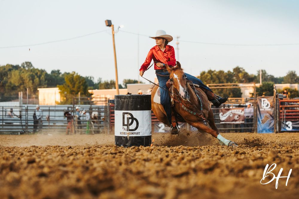 Barrel racer on horseback rounding a barrel at a rodeo competition in a dirt arena.