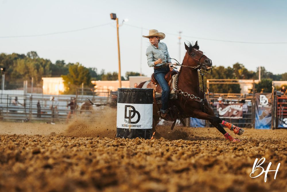 Barrel racer on a chestnut horse competing in a rodeo arena, turning around a barrel at high speed.