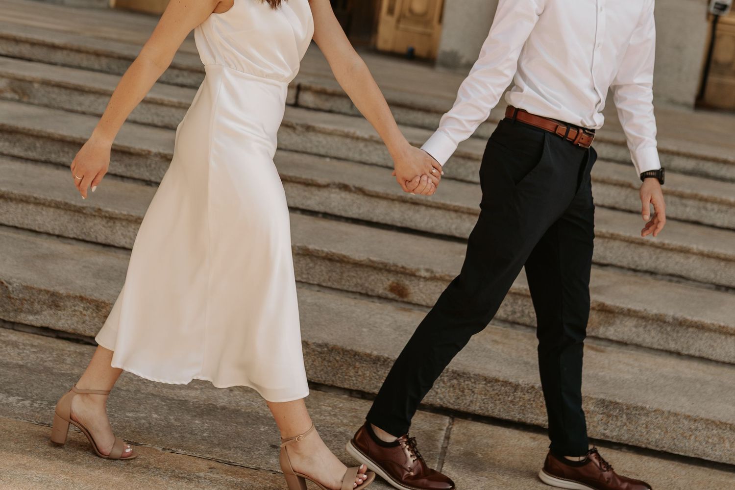 Couple in wedding attire walking hand in hand through a sunlit forest meadow at golden hour.