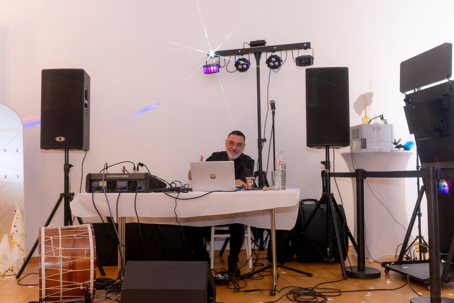 DJ performing at a white studio event setup with speakers, laptop, and stage lighting equipment.