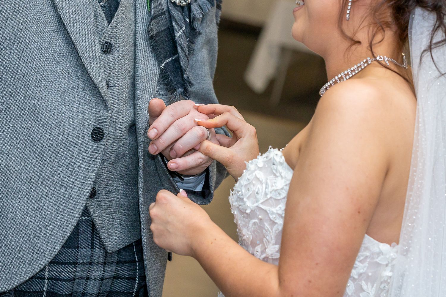 Bride in white lace gown placing wedding ring on groom's finger during ceremony, groom wearing gray tartan kilt suit.