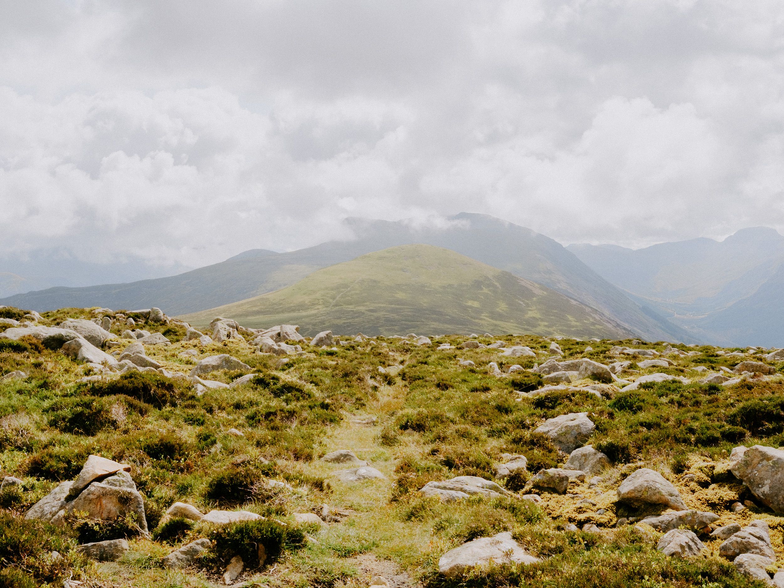 Rocky mountain trail leading through a rugged highland landscape with green moss, boulders, and misty peaks under cloudy skies.