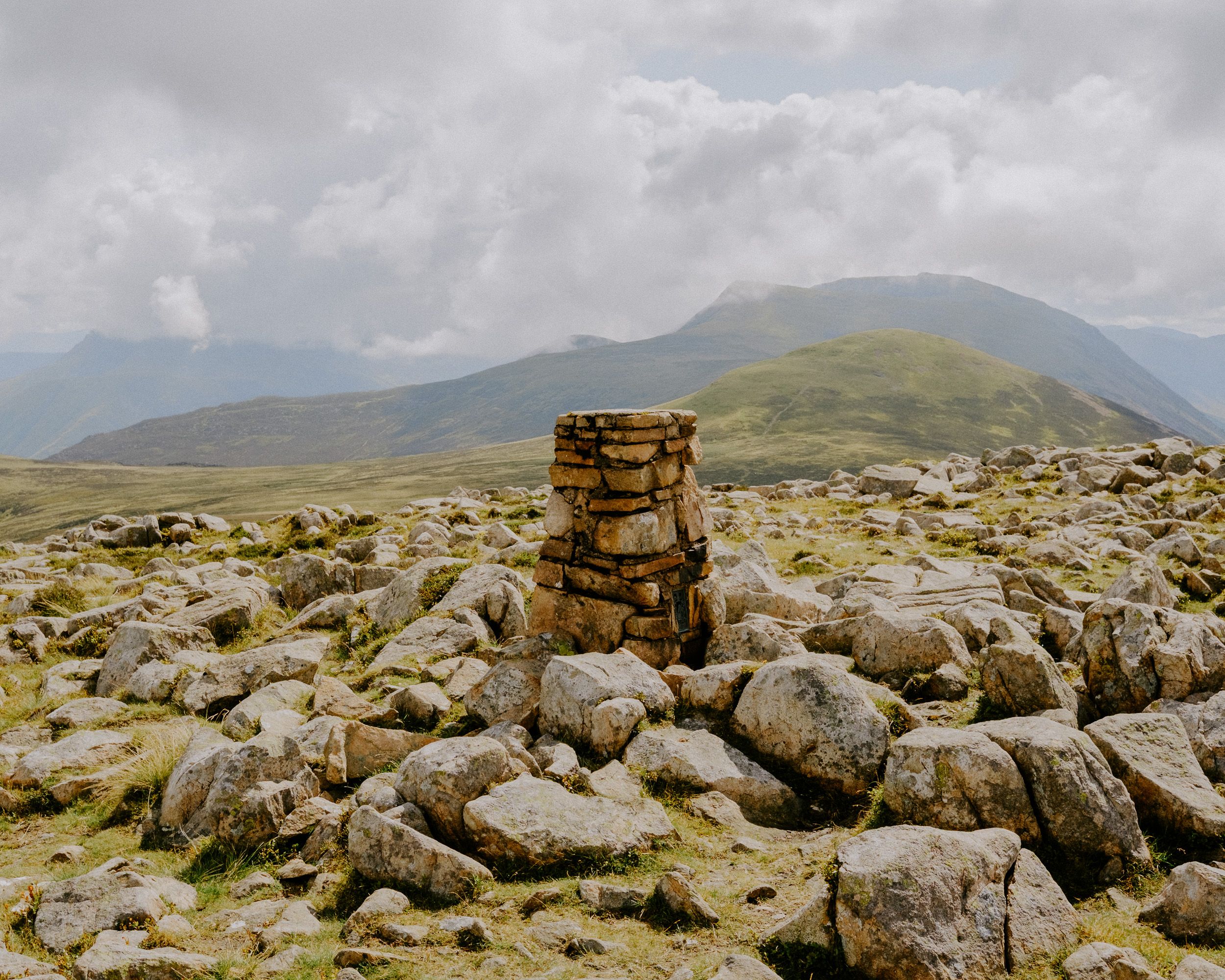 A stone cairn stands among rocky terrain with green hills and cloudy mountain landscape in the background.