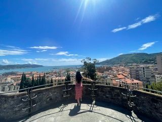 Woman standing on a stone viewpoint overlooking a coastal city with red rooftops, mountains, and blue sky in Europe.