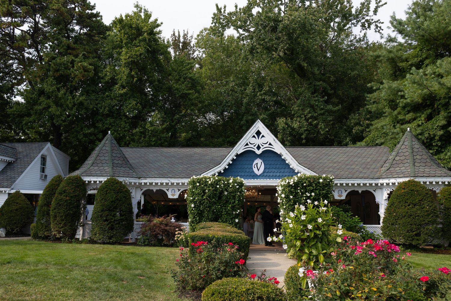 Elegant white wedding venue with blue gabled entrance, lush hedges, and colorful roses in a garden setting.