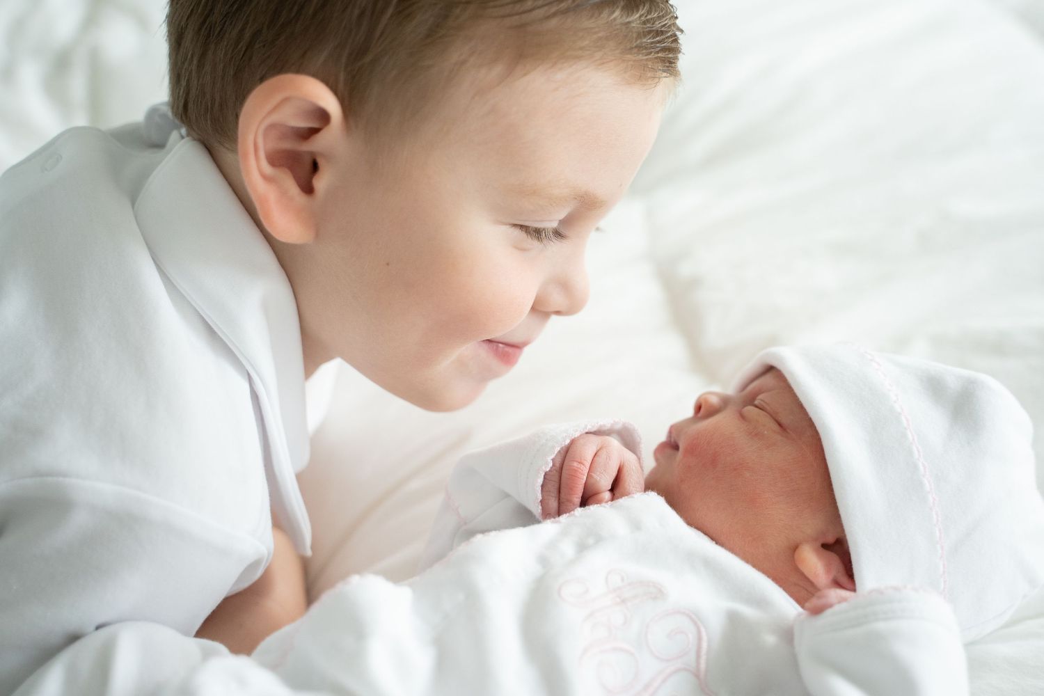 Young boy gazing lovingly at newborn baby sibling wrapped in white blanket on white bed.