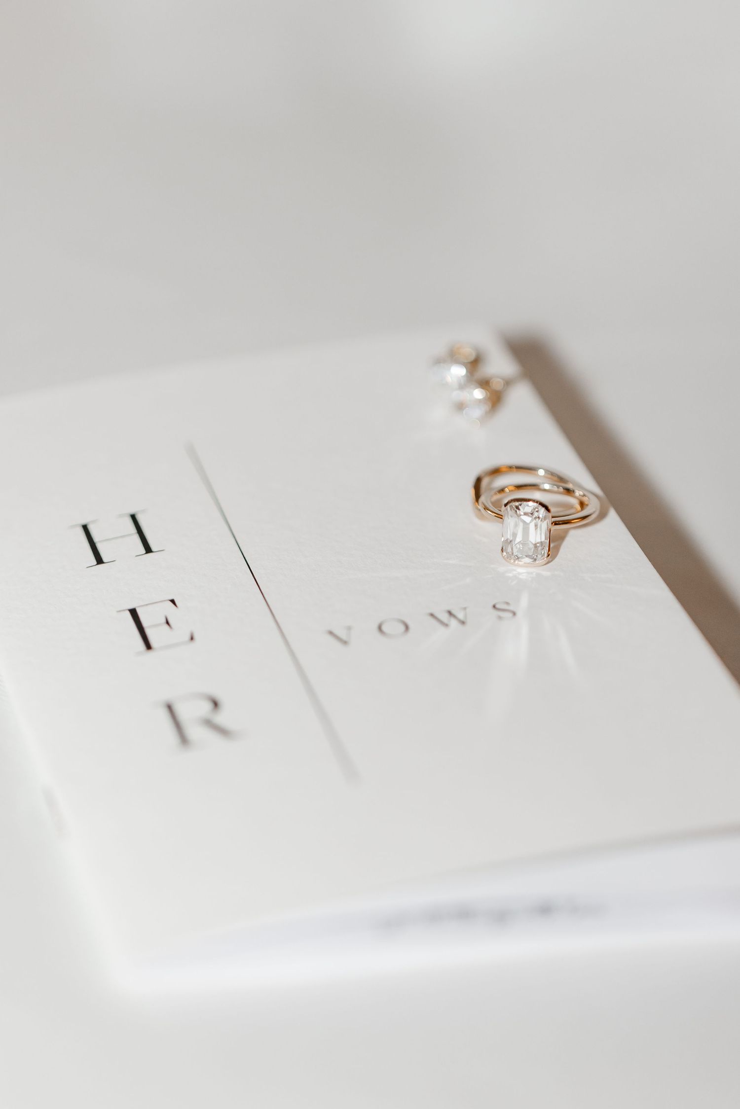 Gold diamond engagement ring and wedding band on a white HER vows book with pearl earrings.