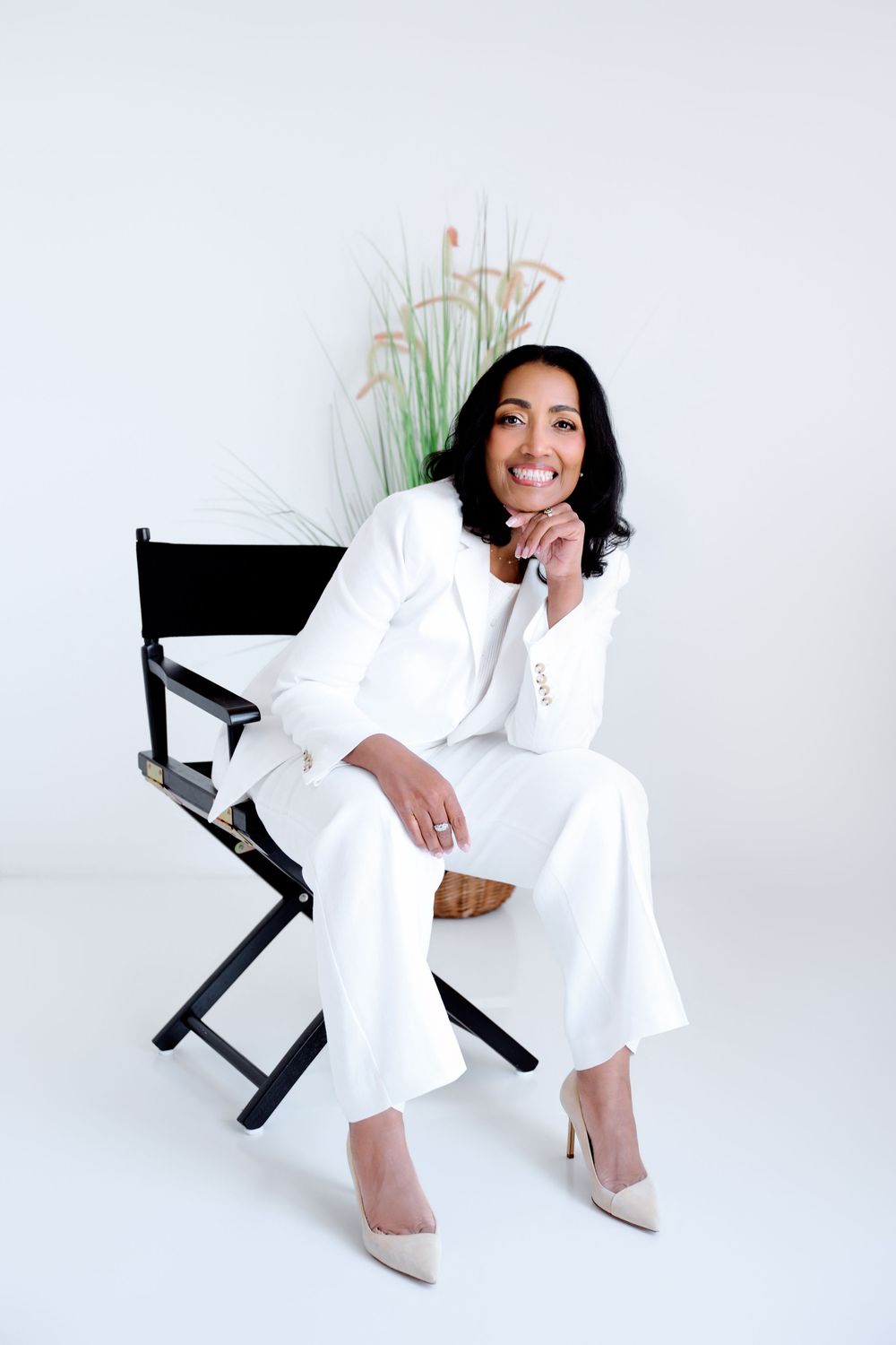 Woman in white suit sitting on director's chair, smiling confidently against white background with decorative grass plant.