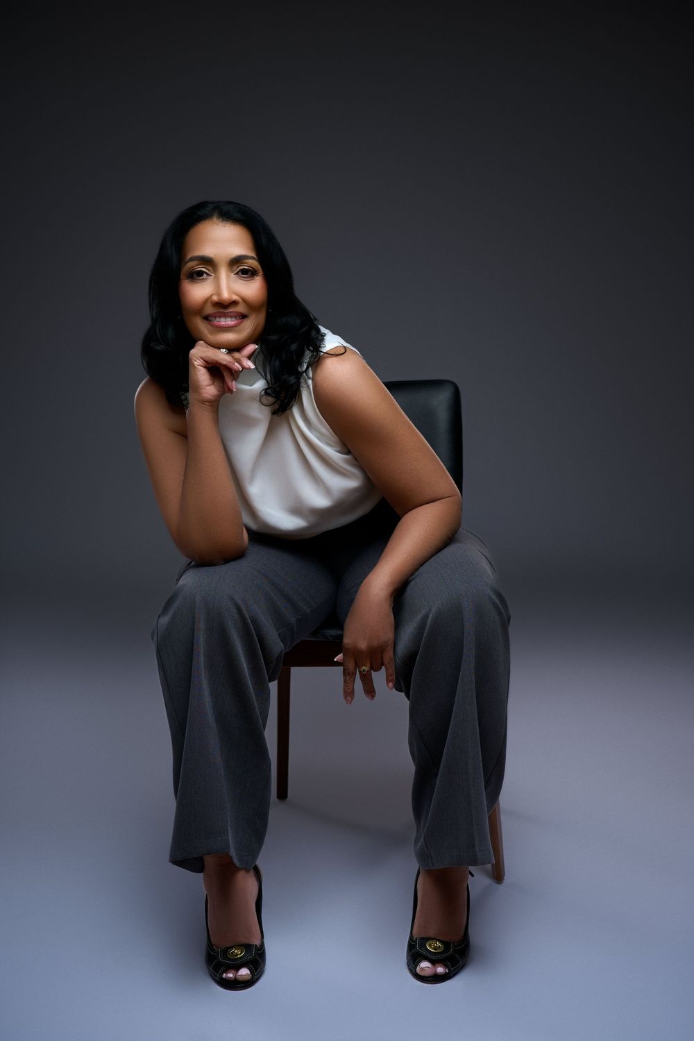A confident woman in a white blouse and gray trousers sits on a chair, smiling with hand on chin against a gray backdrop.