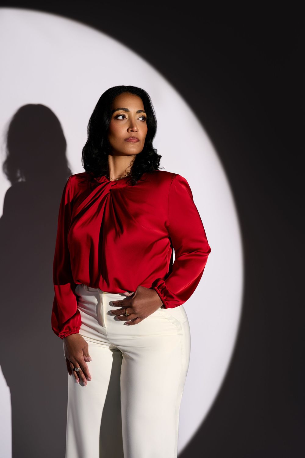 Woman in red satin blouse and white pants posing against dark background with circular spotlight.