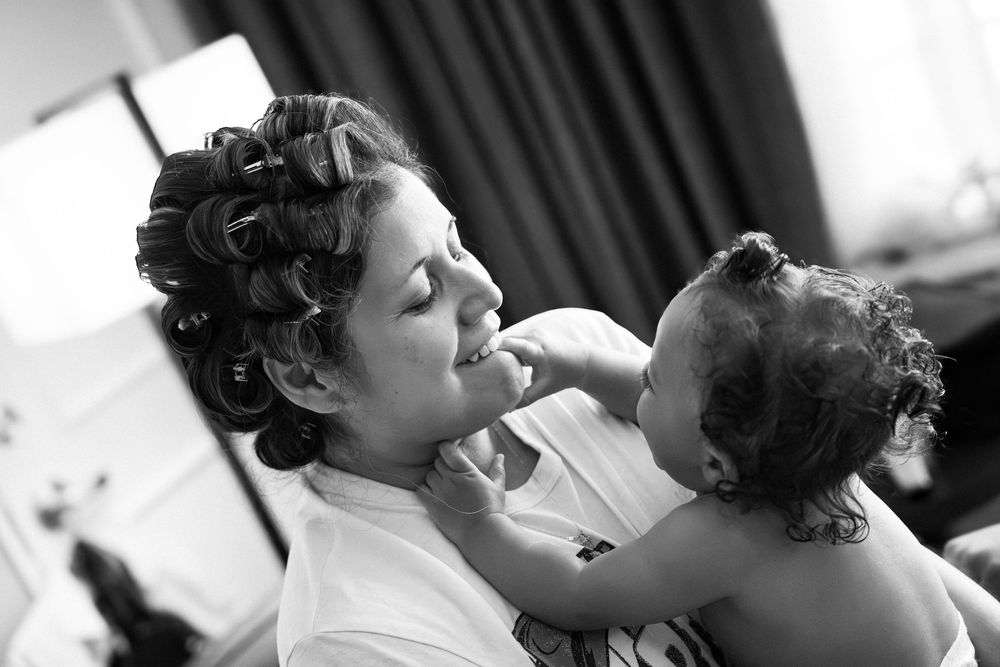 Bride with curly updo holds smiling baby during wedding preparations in black and white photo.