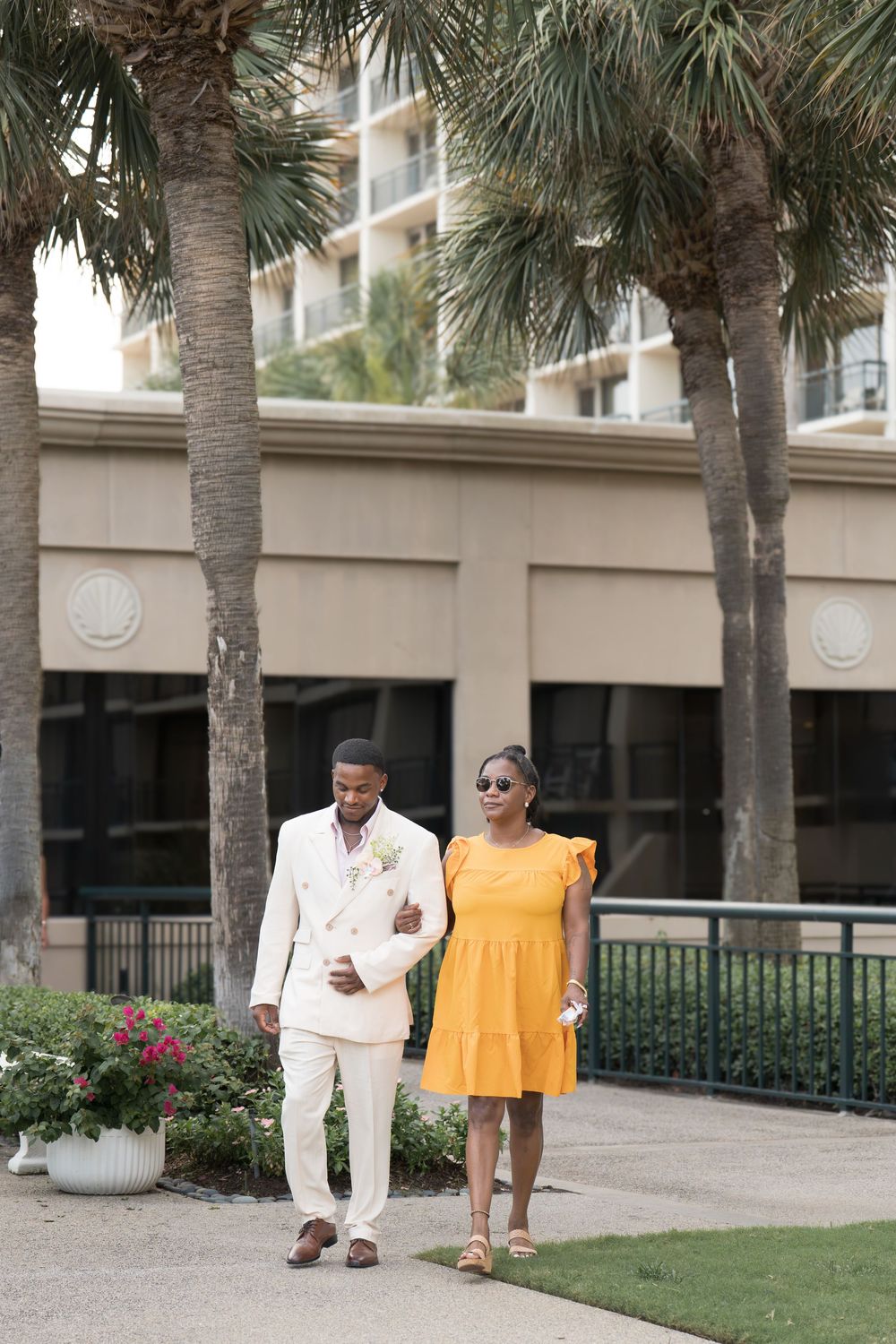 Groom in cream suit walks with woman in yellow dress near palm trees.